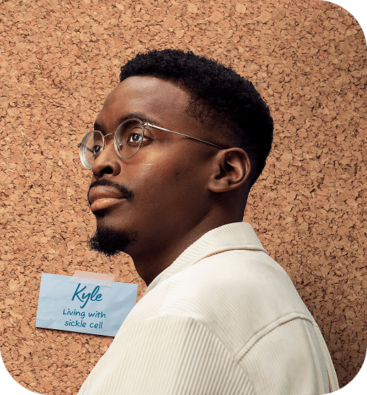 Portrait of a man wearing glasses and a ‘Living with sickle cell’ name badge taped to a corkboard