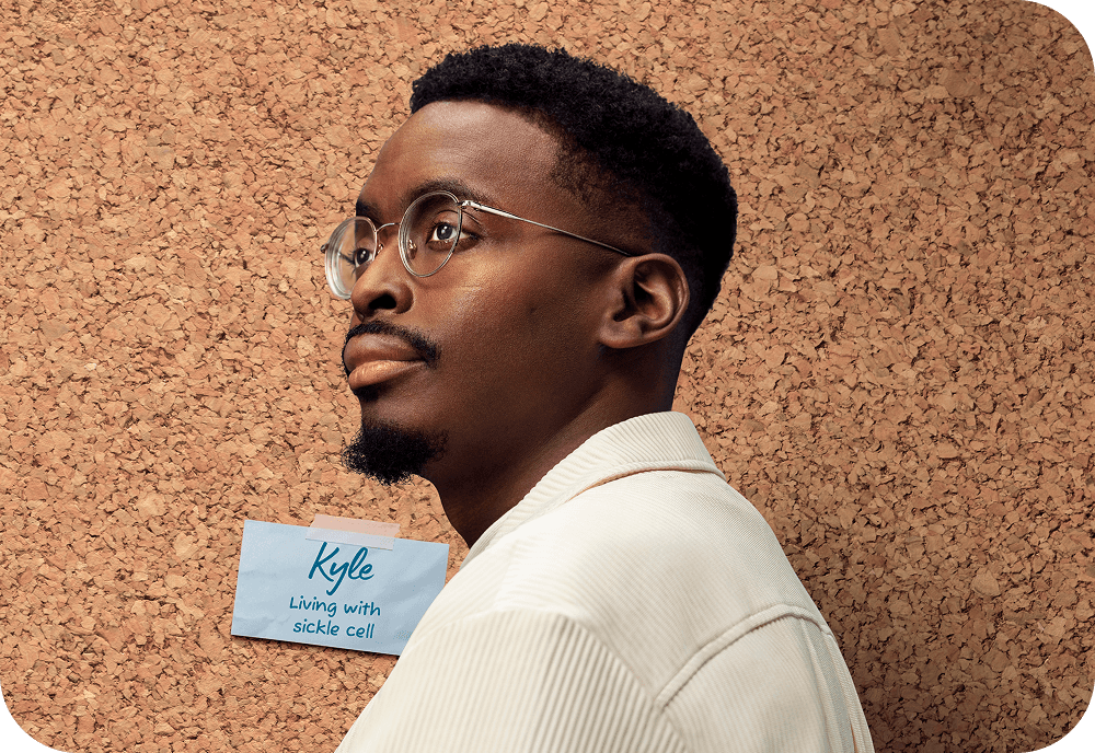 Portrait of a man wearing glasses and a ‘Living with sickle cell’ name badge taped to a corkboard