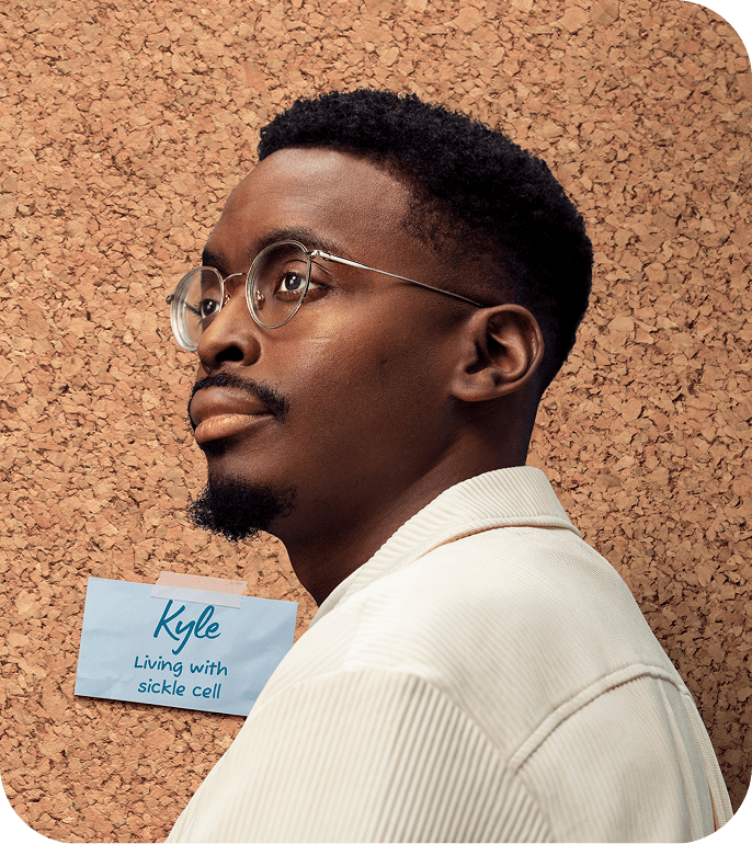 Portrait of a man wearing glasses and a ‘Living with sickle cell’ name badge taped to a corkboard