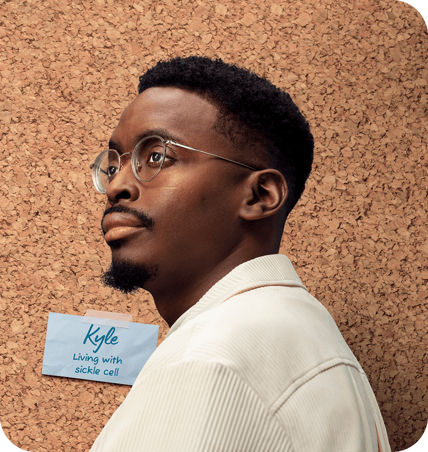 Portrait of a man wearing glasses and a ‘Living with sickle cell’ name badge taped to a corkboard